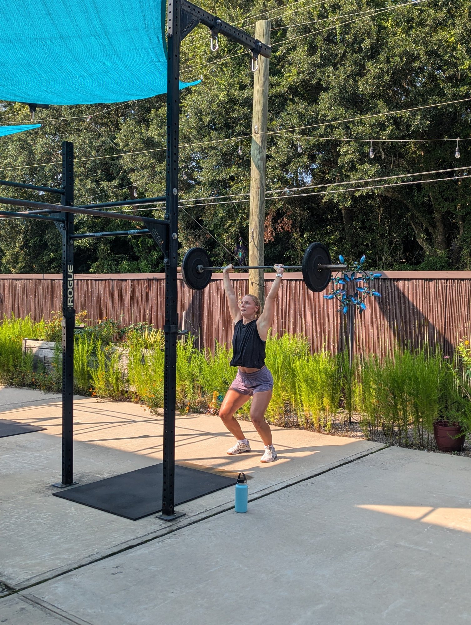 A member lifting a barbell beneath the teal shade sails in Nomad Fitness’s outdoor training space